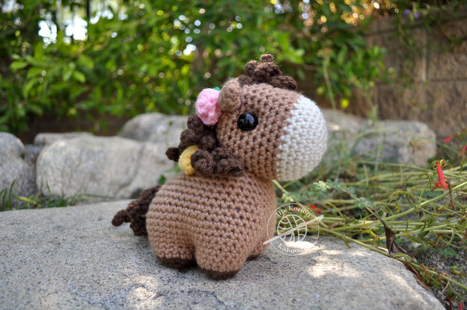 Crocheted horse toy with a pink flower on a stone surface with greenery in the background